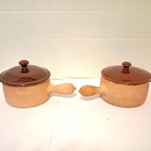 May include: Two small, tan ceramic pots with lids. Each pot has a brown lid and a matching handle. The lids have a small, round knob on top. The pots are likely for cooking or serving.