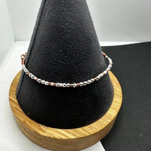 May include: A silver and copper beaded bracelet with a delicate design. The bracelet is displayed on a wooden stand.