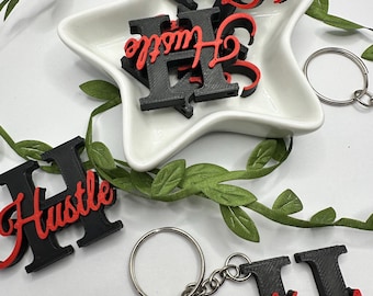 May include: Black and red letter H keychain with the word "Hustle" written in red cursive. The keychain is on a white star-shaped dish with green leaves in the background.