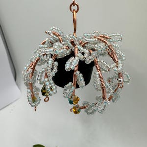 May include: A hanging planter with a black pot, adorned with clear and blue beads strung on copper wire. The beaded strands cascade downwards, with a few yellow and teal beads adding color. The planter is suspended by a copper hook.