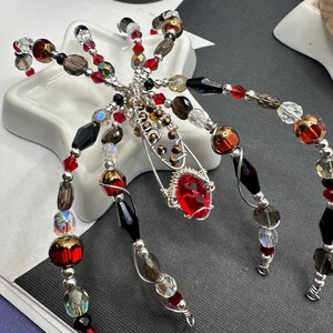 May include: A silver wire spider with red, black, brown, and clear glass beads. The spider has a large red gemstone in the center.