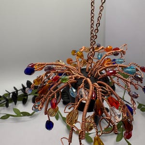 May include: A handcrafted copper wire tree sculpture with colorful crystal beads. The tree is suspended by a copper chain. The beads are various colors, including blue, red, yellow, and clear. The sculpture is a decorative item.