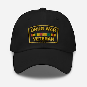 Puede incluir: Gorra de béisbol negra con un parche bordado que dice "Drug War Veteran" con un borde amarillo y una cinta roja, verde y amarilla.