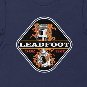 May include: Navy blue t-shirt featuring a diamond-shaped graphic with a piston design engulfed in flames. The graphic includes the text "LEADFOOT 502 HEY NOW!" in white and orange. The shirt is made of a soft, comfortable material.