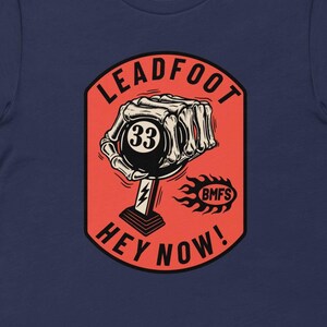 May include: Navy blue t-shirt featuring a graphic with the text "LEADFOOT HEY NOW!" and a skeleton hand holding a black 8-ball with the number 33. The design is in a red and white color scheme with a flame graphic.