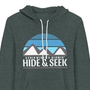 May include: A dark green hooded sweatshirt with a graphic design of a blue and white sunset with mountains and the text "A cold wind is a-blowin' Hide & Seek".