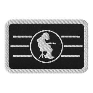 May include: A white embroidered patch with a black background. The patch features a silhouette of a person sitting on a stool, inside a circle. The circle is surrounded by three horizontal lines.
