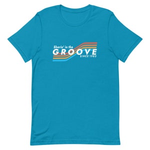 May include: A turquoise blue t-shirt with a retro-style graphic design. The graphic features a rainbow of colors and the text "Sharin' in the GROOVE since 1983".