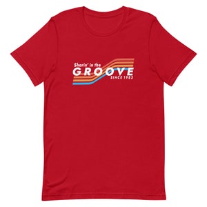 May include: Red t-shirt with a retro-style graphic that says "Sharin' in the GROOVE since 1983". The graphic features colorful stripes.