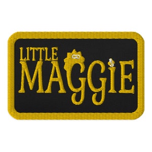 May include: A black and gold embroidered patch with the text "LITTLE MAGGIE" in a stylized font.