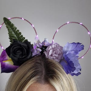 Puede incluir: Una diadema con dos marcos circulares de alambre envueltos en luces LED de color morado. La base está decorada con una rosa negra, un clavel morado claro, un iris morado oscuro, una hoja de helecho verde y una pequeña figura de un personaje.