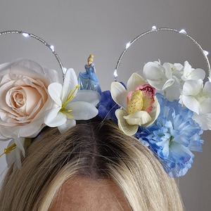 Puede incluir: Una diadema con dos marcos circulares de alambre adornados con pequeñas luces LED blancas. La base está decorada con un arreglo de flores artificiales en tonos rosa pálido, blanco y azul, con una pequeña figura de Cenicienta en el centro.