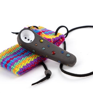 Peut inclure: Une pipe en céramique gris foncé avec des points colorés et un bol blanc. Elle repose sur une pochette tricotée multicolore vibrante et est attachée à un cordon noir. Le design est moderne et artistique.