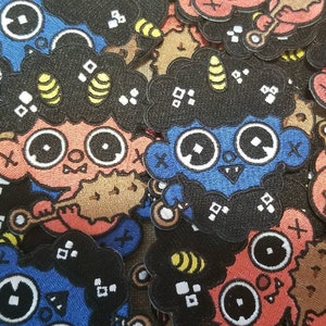 Op de afbeelding: Een verzameling kleurrijke geborduurde patches met cartoonfiguren met zwart haar, blauwe en rode huid en geometrische patronen. De patches zijn gemaakt van vilt en hebben een gestikte rand.