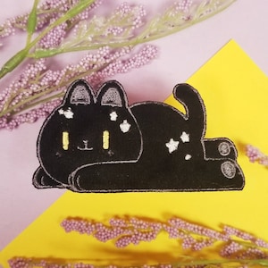 Puede incluir: Parche de hierro bordado negro de un gato con estrellas blancas en su pelaje, acostado de lado con las patas metidas. El gato tiene ojos amarillos y una nariz blanca.