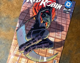Op de afbeelding: Een stripboek getiteld Absolute Red Robin All In nummer 3 met een personage in een zwart pak en vogelmasker dat een grote zeis vasthoudt. De achtergrond toont een rasterachtige gevel van een gebouw met oranje verlichting en een blauw logo.