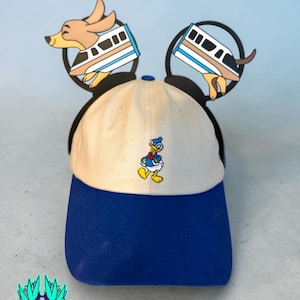 May include: A beige and blue baseball cap featuring an embroidered Donald Duck character on the front. Attached to the cap are black circular ears, each decorated with a graphic of a monorail train and a tan dog character.