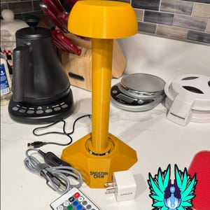 Puede incluir: Una lámpara amarilla con una parte superior en forma de hongo y una base hexagonal. La base tiene el texto "SKELETON CREW". Incluye un mando a distancia, un cable USB y un adaptador de corriente. La lámpara está sobre una superficie blanca.