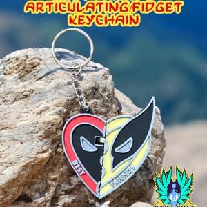 May include: A metal keychain featuring Deadpool and Wolverine's faces on a heart-shaped design. The keychain is articulated and has a silver chain and ring. The text "Best Friends" is printed on the keychain. #GalacticMercantileGuild