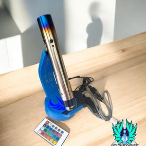 Peut inclure: Un sabre laser argenté et bleu avec un support bleu et une télécommande avec des options de couleurs. Le sabre laser a une pointe aux couleurs de l'arc-en-ciel et repose sur une surface en bois. L'image comprend également un logo avec du texte.
