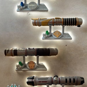 May include: A collection of four lightsaber replicas on display stands. Each lightsaber is a different color and has a unique design. The stands are made of a light gray material and feature a crystal and a plaque with the name of the lightsaber.