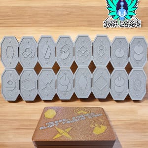 May include: A set of 16 grey octagonal tiles with different symbols on each tile. The tiles are arranged in two rows of eight. The tiles are in front of a brown wooden box with gold glitter and the text "KEEP IT SIMPLE" and "SHIFT 7 QUEENS GAME" on the top.