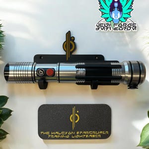 May include: A black lightsaber wall mount with a silver lightsaber attached. The lightsaber has a black handle and a silver blade. The wall mount has a gold logo that says "The Halcyon Starcruiser Training Lightsaber".