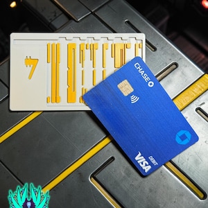 Könnte beinhalten: Eine blaue Chase Visa Debitkarte mit einem Symbol für kontaktloses Bezahlen liegt auf einer metallischen Oberfläche neben einem weißen und gelben rechteckigen Design mit der Zahl 7. Das Bild enthält auch ein Logo mit Text.