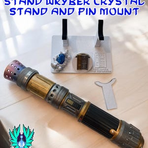以下が含まれることがあります： 黒、金、銀色の柄のライトセーバーが、2つの黒いサポートが付いた白いスタンドに置かれています。スタンドには青いクリスタルと小さなドアのデザインもあります。テキストには「RECLAIMED AND REFORGED LIGHTSABER STAND WKYBER CRYSTAL STAND AND PIN MOUNT」と書かれています。