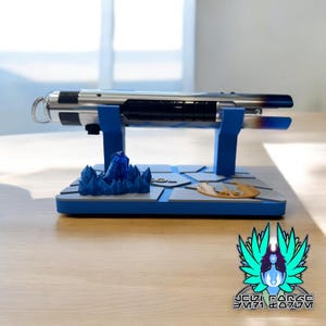 May include: A blue and silver lightsaber on a blue and gray display stand with a blue crystal and a gold symbol. The stand is on a wooden surface.