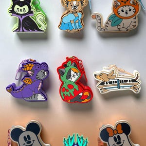May include: A collection of eight colorful silicone keychains featuring various cartoon characters, including a cat dressed as a villain, a dog in a blue dress, a cat in an orange hat, a purple dragon, a cat in a red hood, a dog on a monorail, and two ghost-shaped characters, with a logo at the bottom.