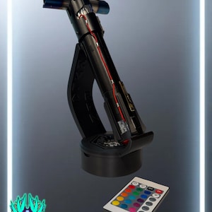 May include: A black crossguard lightsaber hilt with exposed red wiring and blue-tinted emitter tips, mounted on a curved black display stand. A white remote control with multiple colored buttons for light adjustment is positioned in the foreground.
