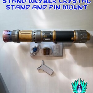 以下が含まれることがあります： 黒、金、銀色の柄のライトセーバーが白いスタンドに置かれています。スタンドには青いクリスタルとピンマウントも含まれています。上部のテキストには「RECLAIMED AND REFORGED LIGHTSABER STAND WKYBER CRYSTAL STAND AND PIN MOUNT」と書かれています。