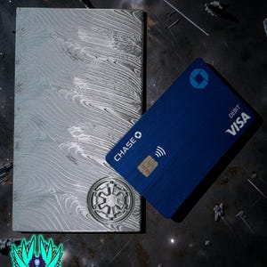 May include: A rectangular silver-colored metal bar featuring a swirling Damascus-style pattern and a circular Galactic Empire emblem embossed in the corner, positioned next to a blue Chase Visa debit card with a chip and contactless payment symbol.