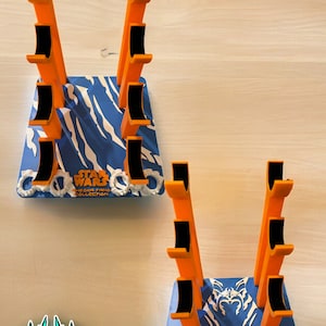 May include: A 3D printed stand for displaying Star Wars Ahsoka Tano figures. The stand is blue and white with a tiger stripe pattern and the words "Star Wars Ahsoka Tano Collection". The stand has six slots for figures and is made of orange and black plastic.