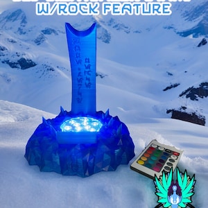 Op de afbeelding: Een blauwe lightsaber-standaard met een rotsfunctie en een afstandsbediening om de kleur van de LED-verlichting te veranderen. De standaard staat op een besneeuwde ondergrond. #GALACTICMERCANTILEGUILD