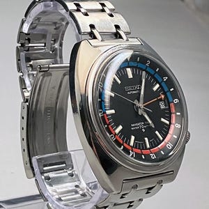 Puede incluir: Un reloj Seiko Navigator Timer vintage con esfera negra, marcadores de hora y agujas blancas. El reloj tiene una correa y caja de acero inoxidable, con detalles azules y rojos en la esfera. La esfera del reloj muestra el texto "SEIKO AUTOMATIC" y "NAVIGATOR TIMER WATER 70m RESIST".