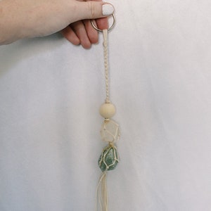 May include: A white string keychain with a wooden bead, a clear crystal, and a green crystal. The crystals are wrapped in macrame.