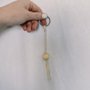 May include: A beige macrame keychain with a wooden bead and a silver key ring.