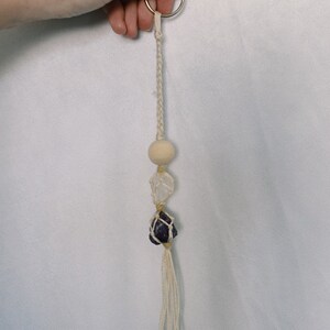 Może przedstawiać: Biały brelok z makramą z drewnianym koralikiem, kryształem górskim i fioletowym ametystem.