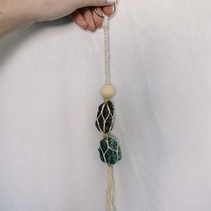 Może przedstawiać: Biały brelok z makramą z drewnianym koralikiem i dwoma kryształowymi kamieniami, jednym ciemnobrązowym i jednym niebiesko-zielonym.