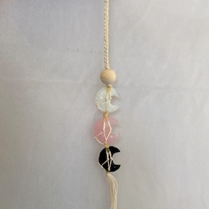 May include: A white, pink and black moonstone keychain with a wooden bead and a white tassel. The keychain is made of macrame and is attached to a silver key ring.