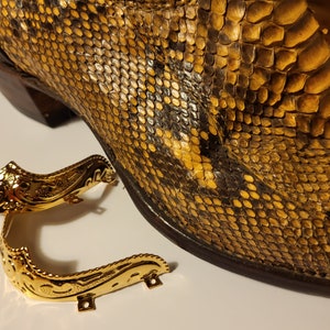 May include: Two gold metal boot heel protectors with ornate designs. The protectors are lying on a white surface next to a brown snakeskin cowboy boot.