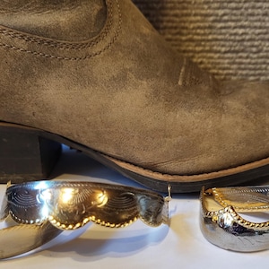 May include: Two metallic boot tips featuring decorative engraved patterns and scalloped edges. One boot tip has a polished gold-tone finish with intricate line work, while the other has a silver-tone finish with a textured, rope-like border design.