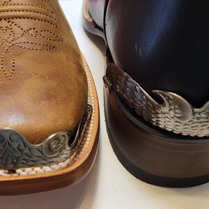 Pu&ograve; includere: Primo piano di due stivali da cowboy in pelle marrone con decorazioni in metallo argentato sulle dita. Gli stivali hanno una punta a punta e un design cucito sulla pelle.