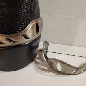May include: A black leather hat with a silver-colored decorative band featuring a wave-like design. A matching silver band is separate from the hat. The hat has a textured surface.