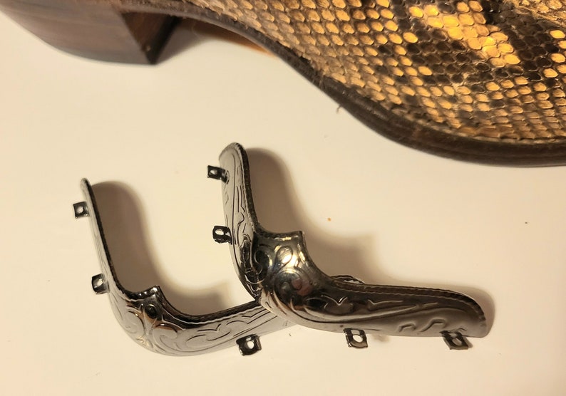 Op de afbeelding: Twee zilveren metalen laarsneuskappen met decoratieve ontwerpen. De kappen hebben kleine gaatjes om aan laarzen te bevestigen.