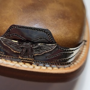 Peut inclure: Gros plan sur une botte en cuir marron avec un motif d'aigle argenté sur la pointe. L'aigle est en relief sur une bande de métal argentée qui entoure la pointe de la botte.