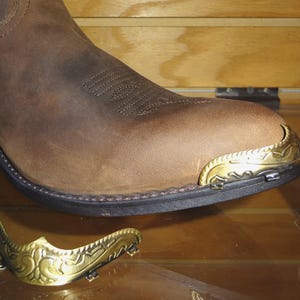 May include: A brown leather boot featuring a decorative gold-toned metal toe cap with an embossed pattern. A matching, separate gold-toned metal toe cap with an intricate scrollwork design is positioned in front of the boot on a surface.