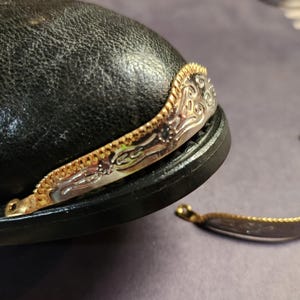 May include: A black leather boot toe featuring an attached silver and gold-toned metal tip with intricate engraved scrollwork patterns. A second matching metal tip is positioned separately to the right, showing the same decorative design.